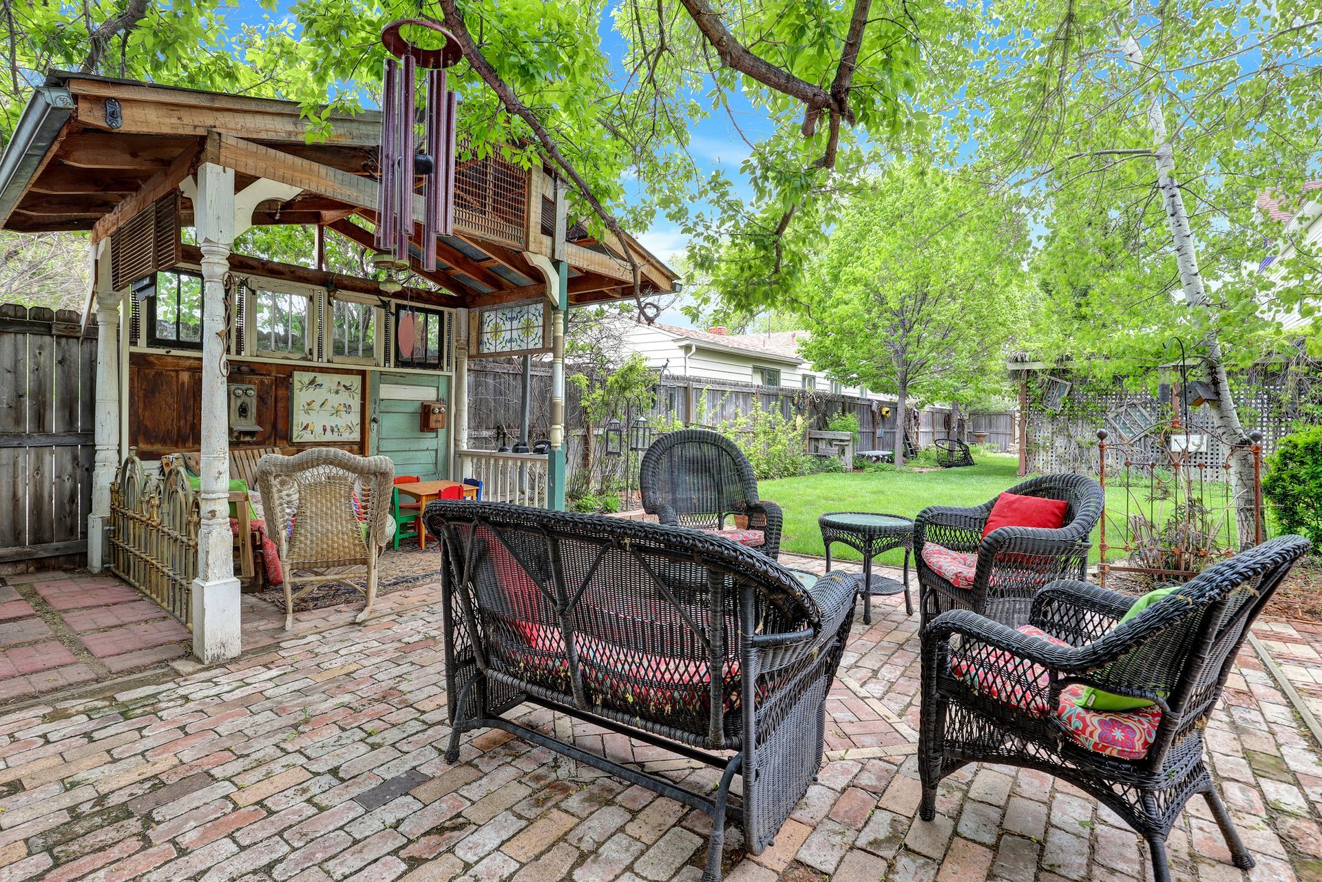 A shaded backyard patio featuring a wooden gazebo with wicker furniture on a brick floor, surrounded by trees and plants.