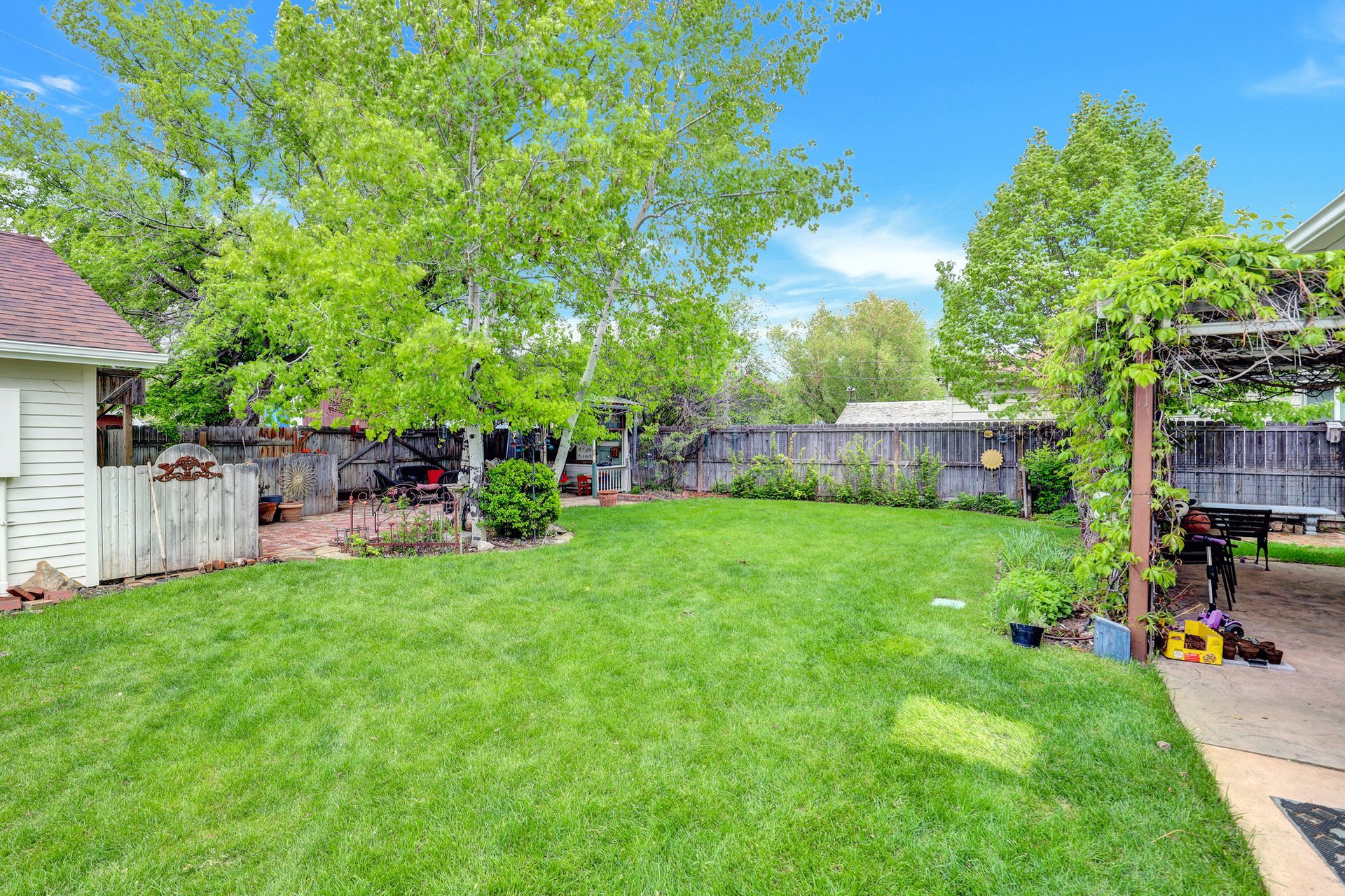 A backyard with a green lawn, lush trees, a wooden fence, and a patio area covered by a vine-covered pergola.