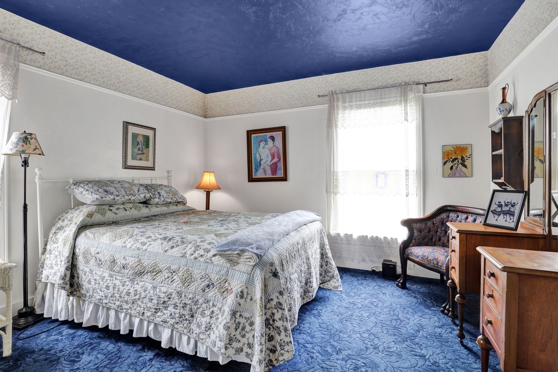 A bedroom with a blue carpet and ceiling, a floral bedspread, a small wood dresser, and an armchair by a bright window.