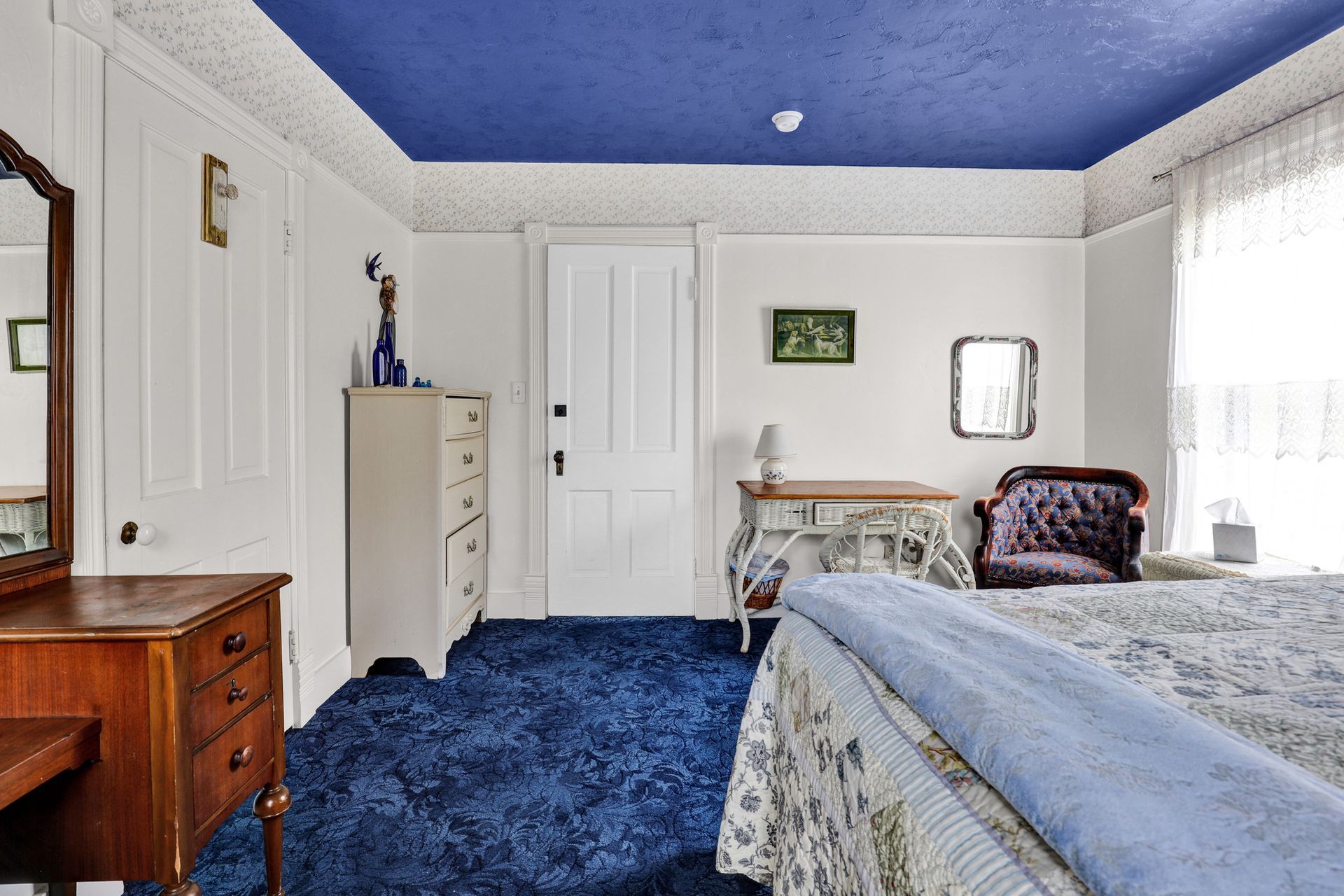 A bedroom with a blue ceiling and carpet, white walls, a wooden vanity, a dresser, a small table, and a patterned chair.