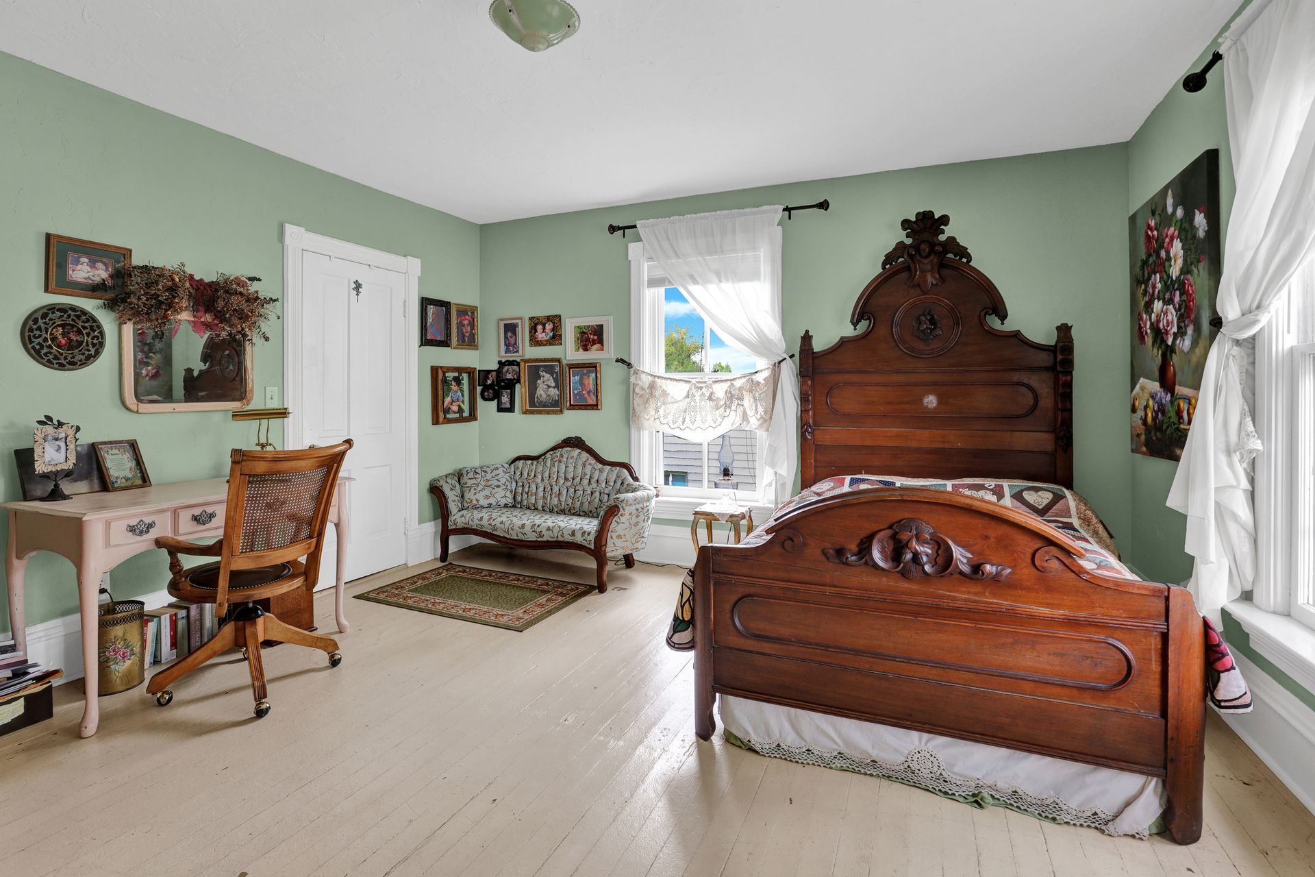 A bedroom with pale green walls, light wood floors, an ornate dark wood bed, a small sofa, and a desk with a chair.
