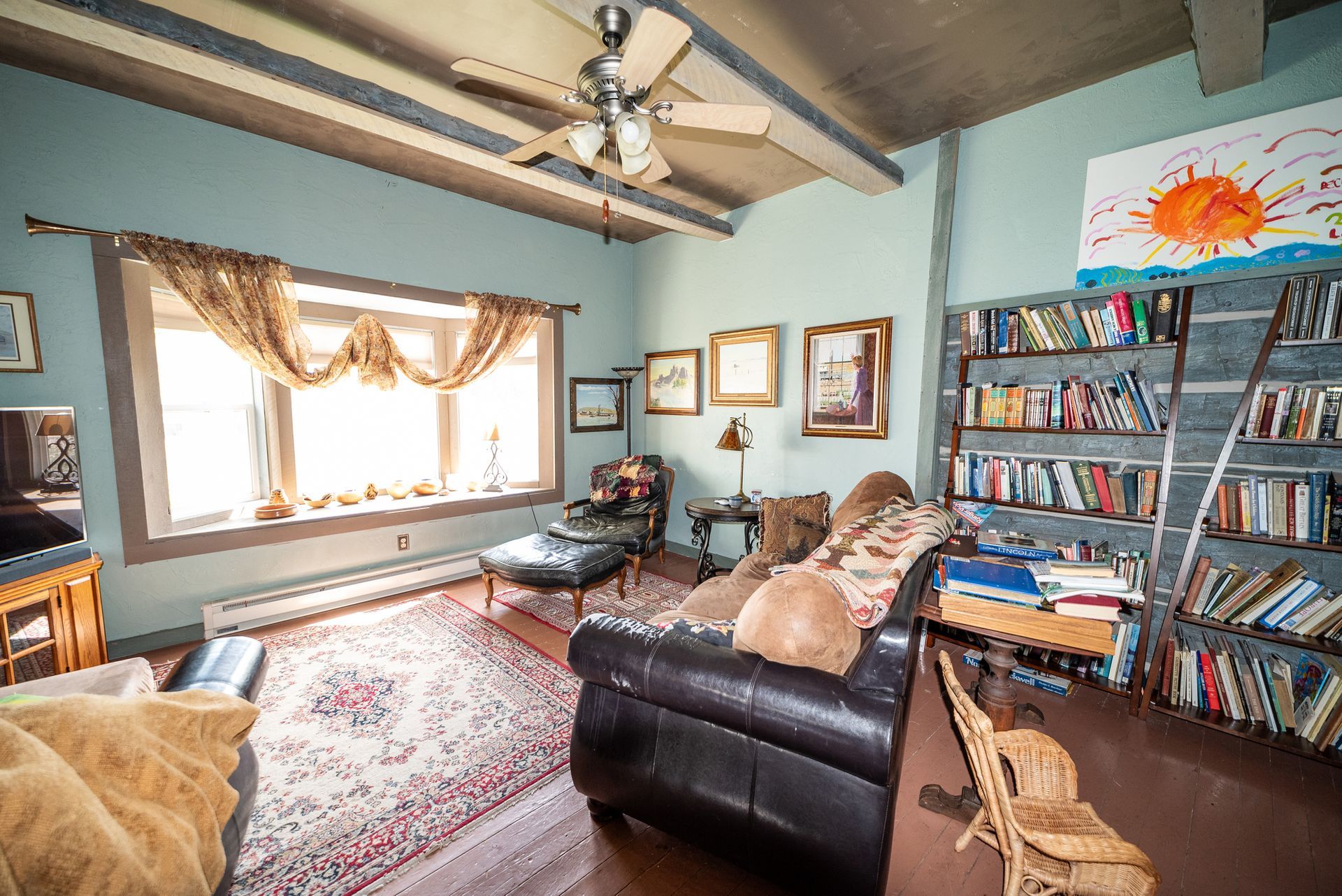 A cozy living room with light blue walls, a dark leather sofa, a patterned rug, and a tall wooden bookshelf filled with books.