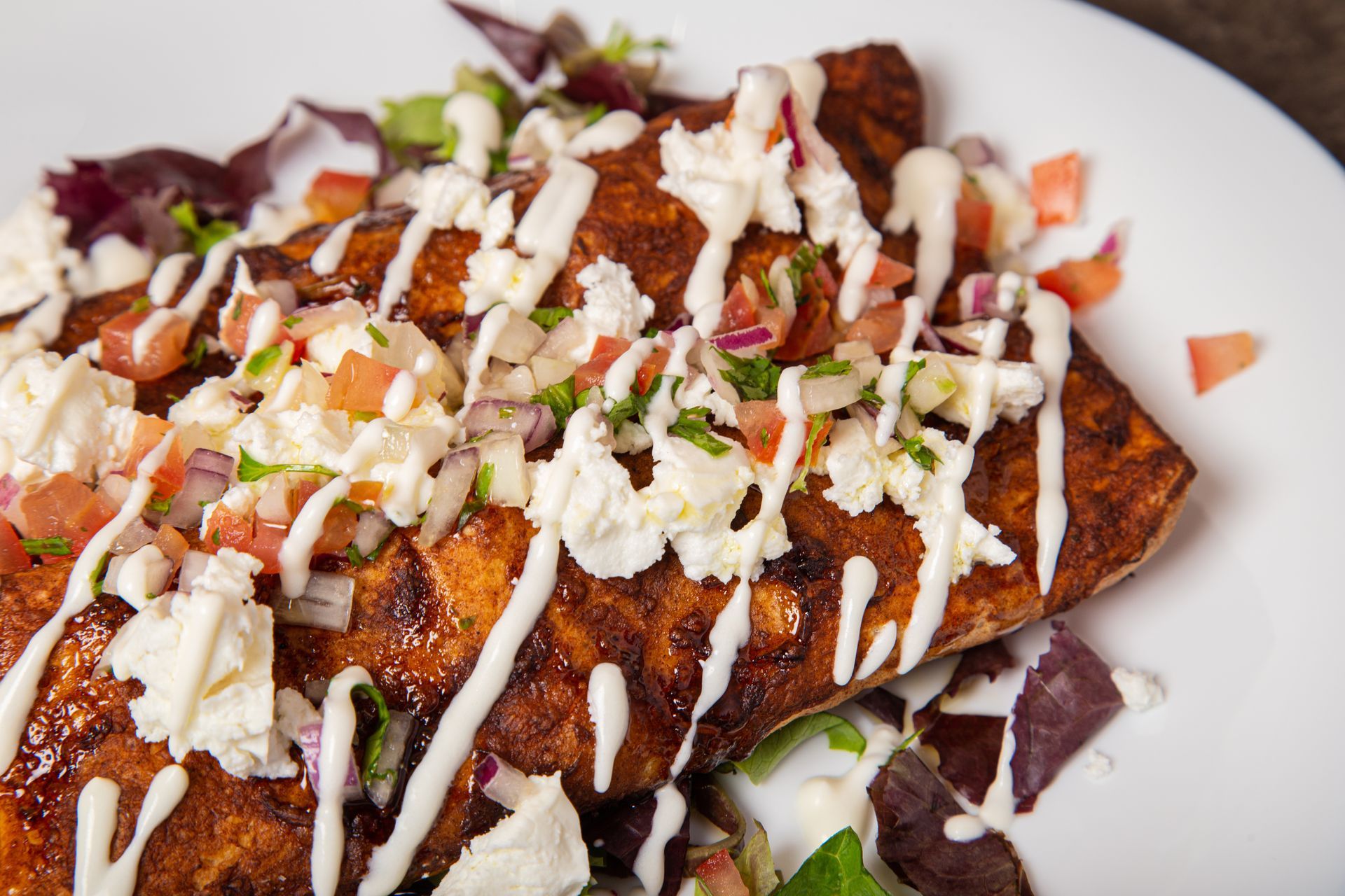 A plate of enchiladas topped with white crema, crumbly cheese, and pico de gallo, resting on a bed of mixed greens.