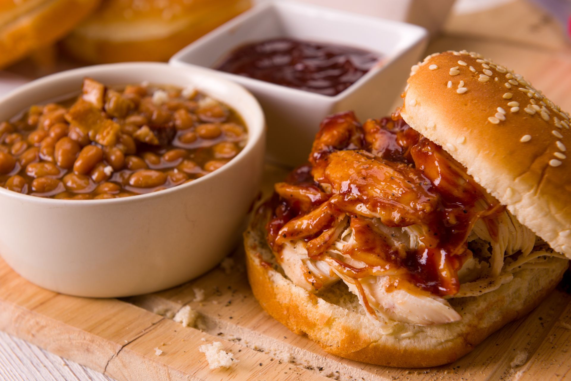 A pulled chicken sandwich with BBQ sauce on a sesame bun, served with a bowl of baked beans and a side of BBQ sauce.