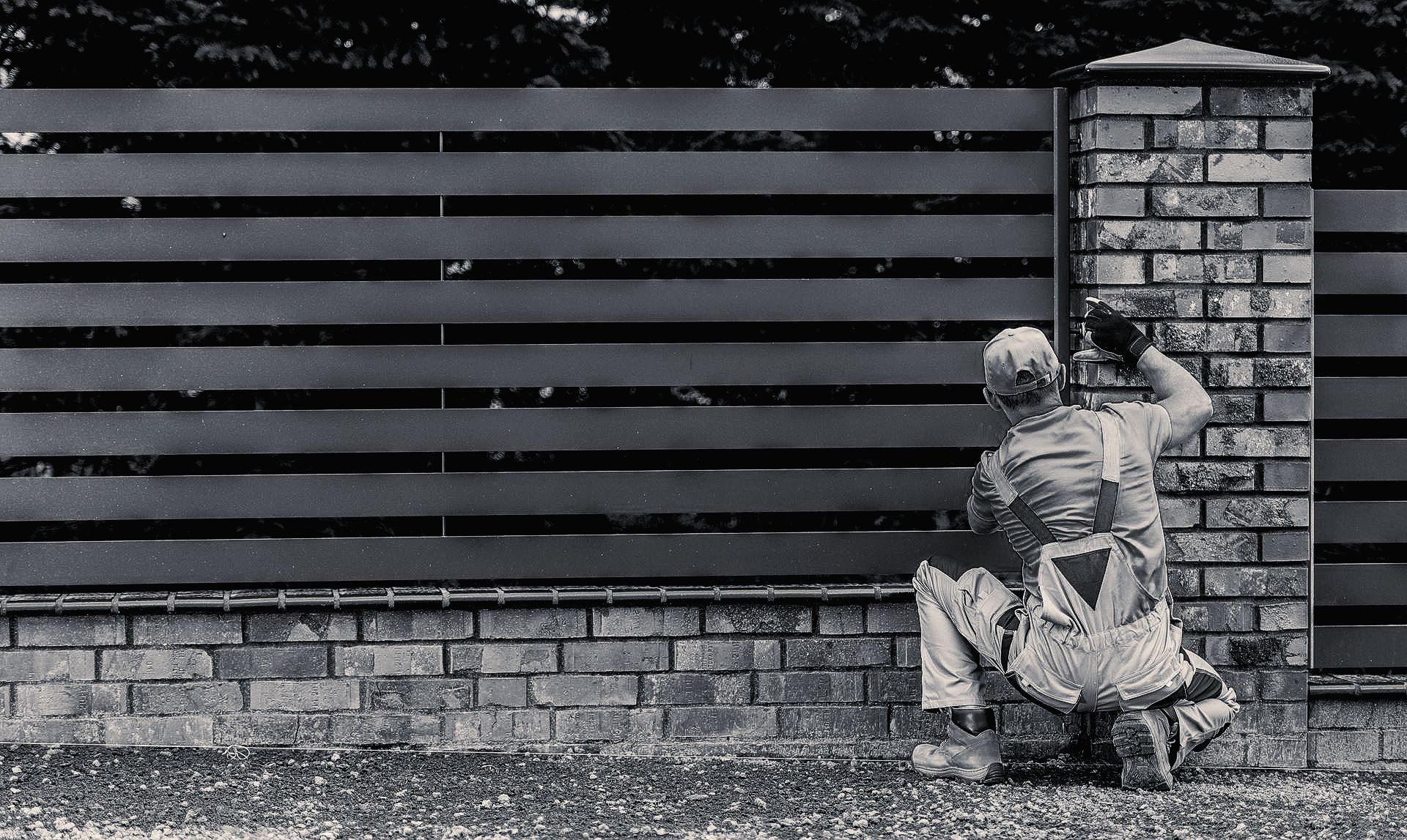 A worker in overalls and hard hat installing or repairing a brick fence post, outdoor setting.