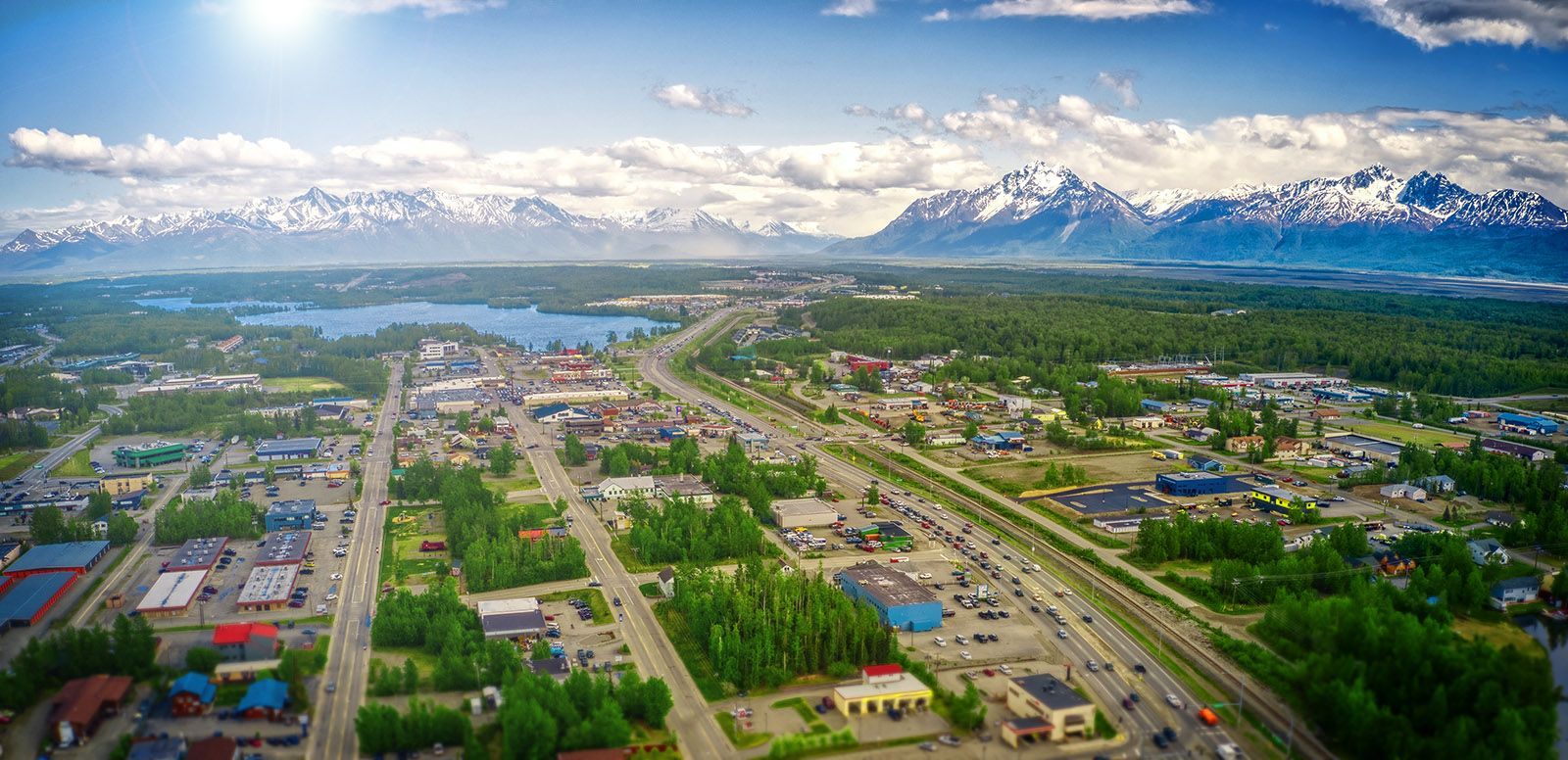 Aerial view of a the Mat-Su Valley with a highway, trees, lake, and snow-capped mountains under a blue sky.