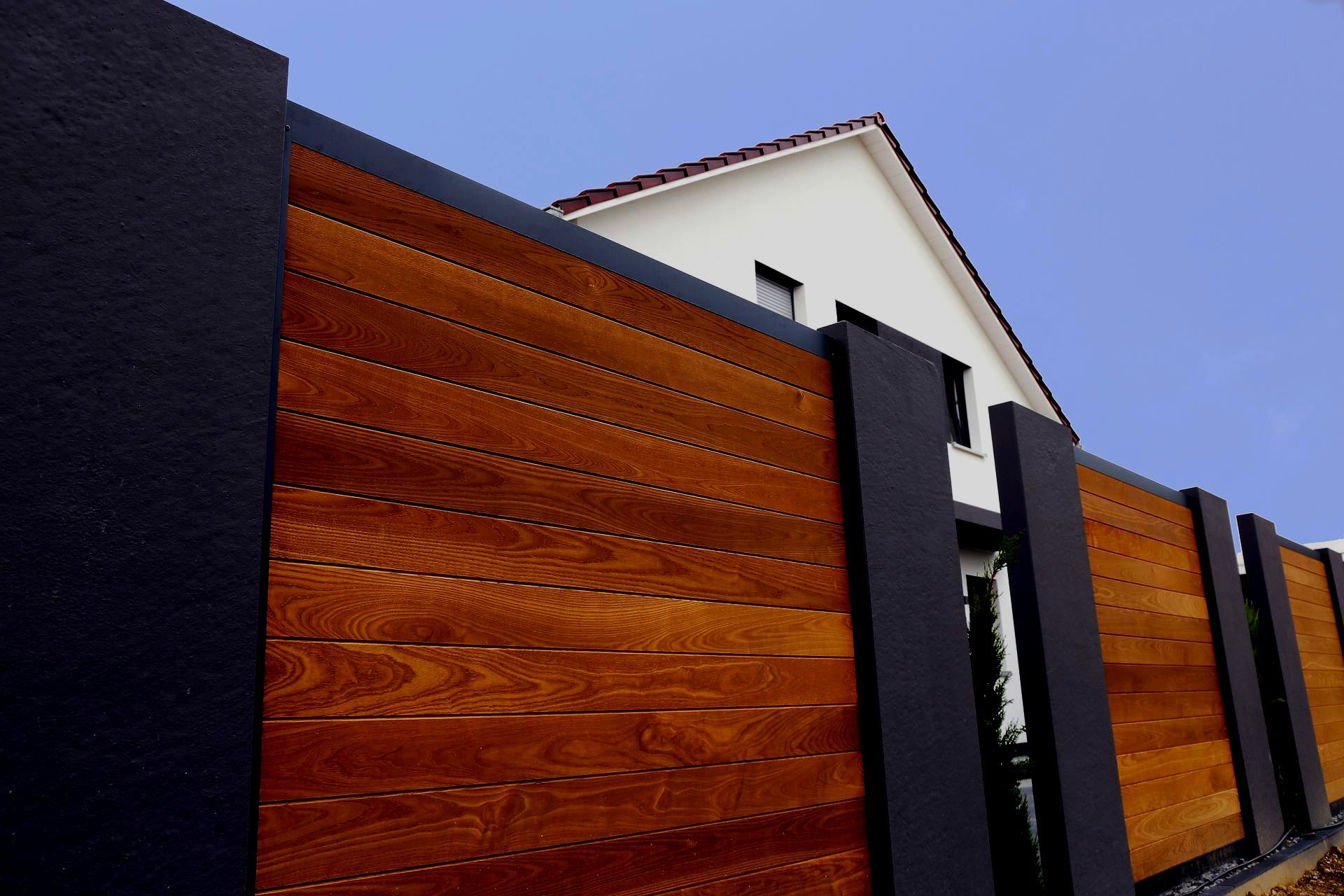 Wooden fence with dark gray pillars, a white house with a red roof is in the background.