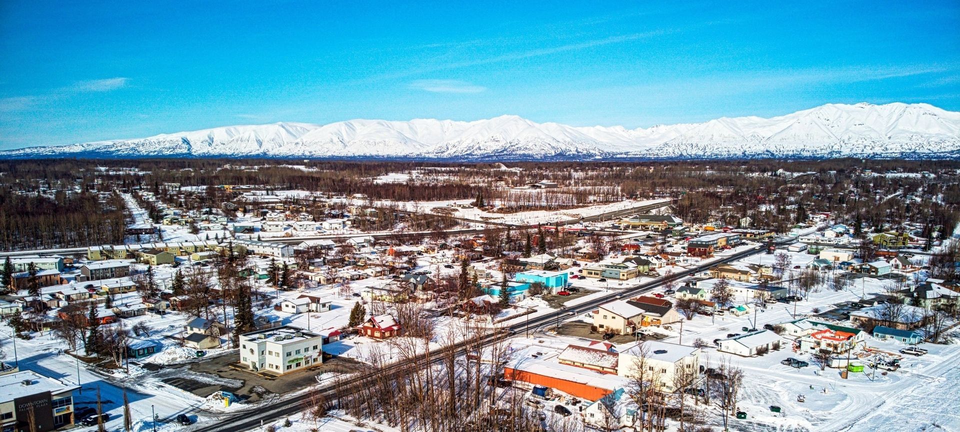 Aerial image of Palmer, AK. Snow-covered town beneath snow-capped mountains, clear blue sky.