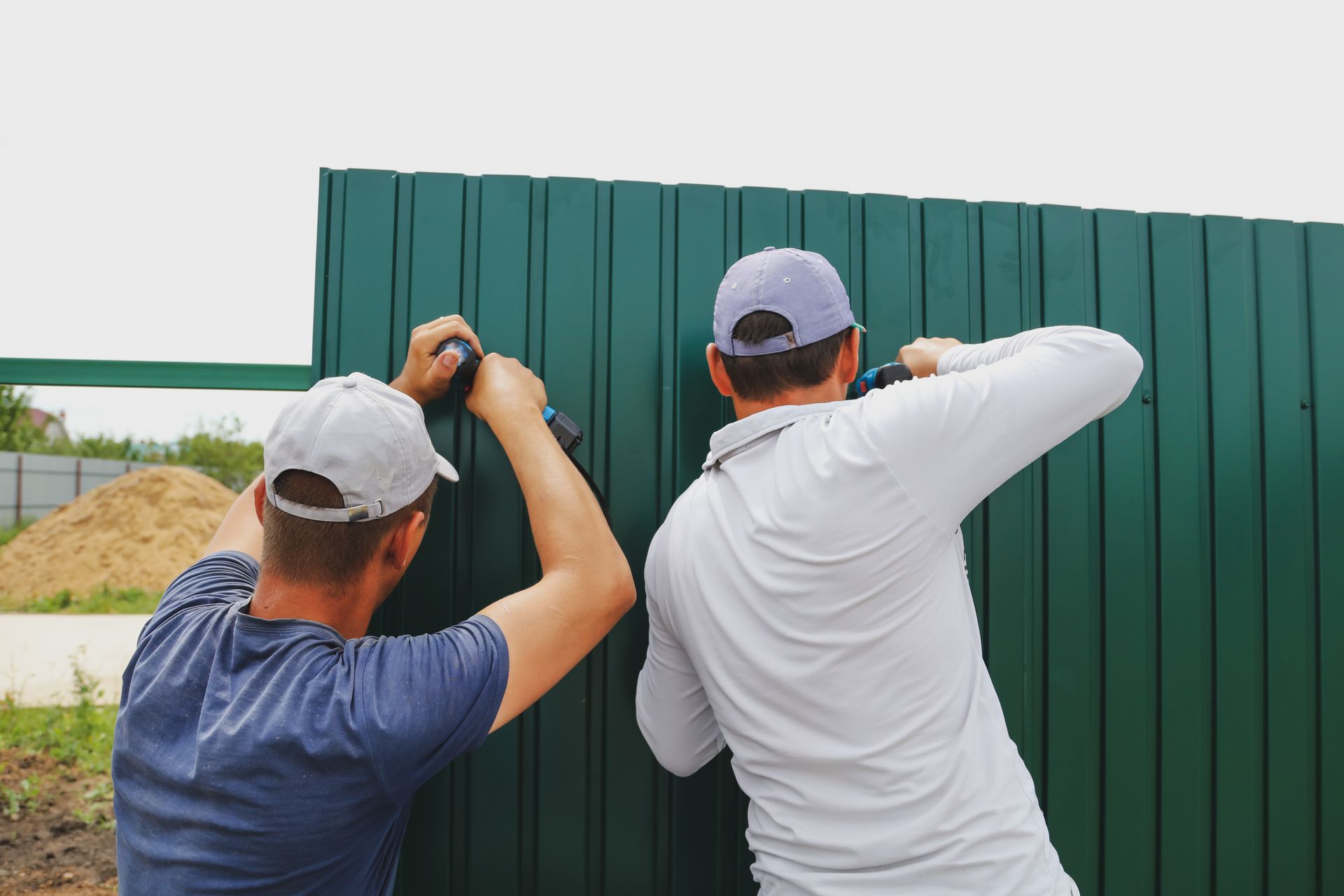Two men in caps installing a green metal fence, using tools.