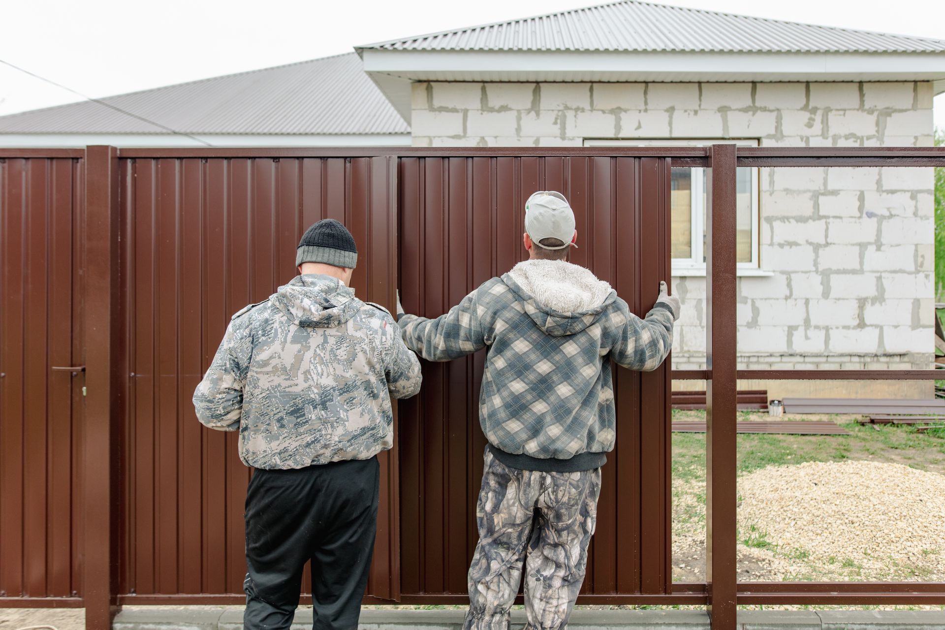 Two people installing a brown metal fence in front of a house.