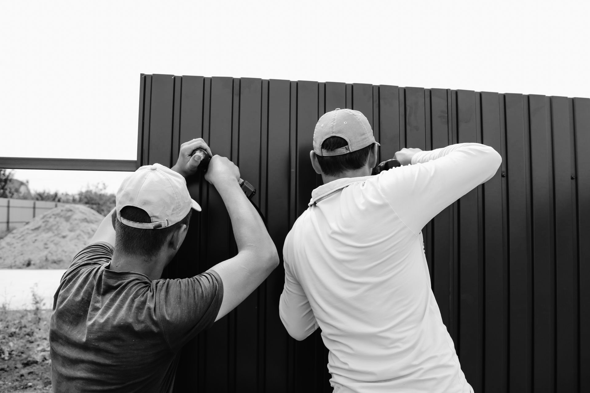 Two men in hats installing a metal fence using power tools outdoors.