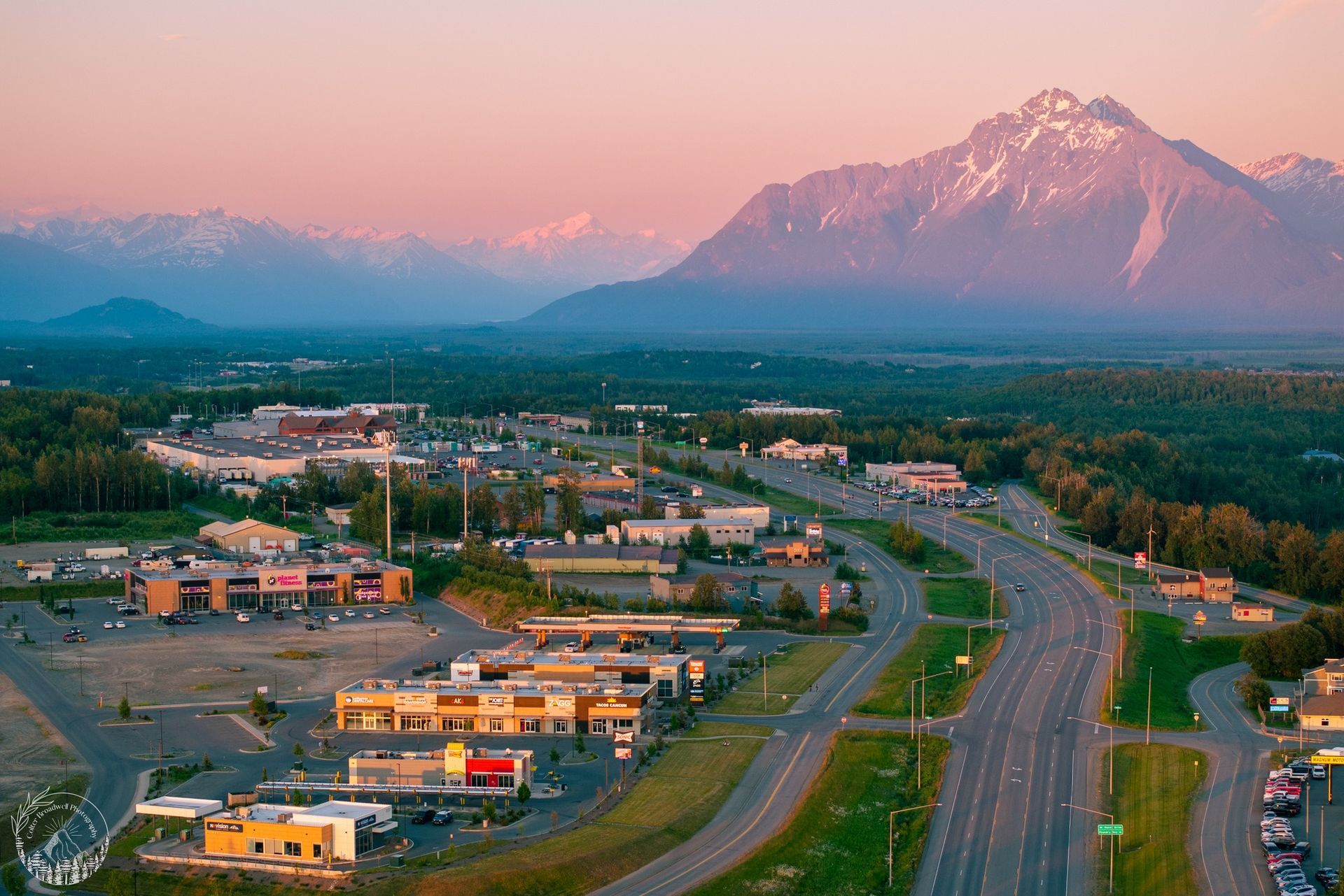 Wasilla, AK aerial image.  Nestled at base of snow-capped mountains; a highway curves through the town.