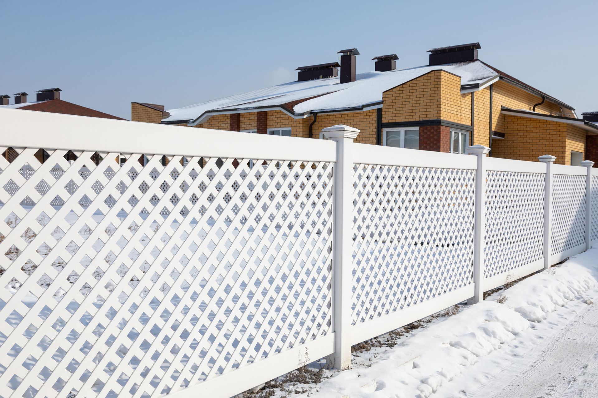 A white lattice fence is surrounded by snow in front of a house.
