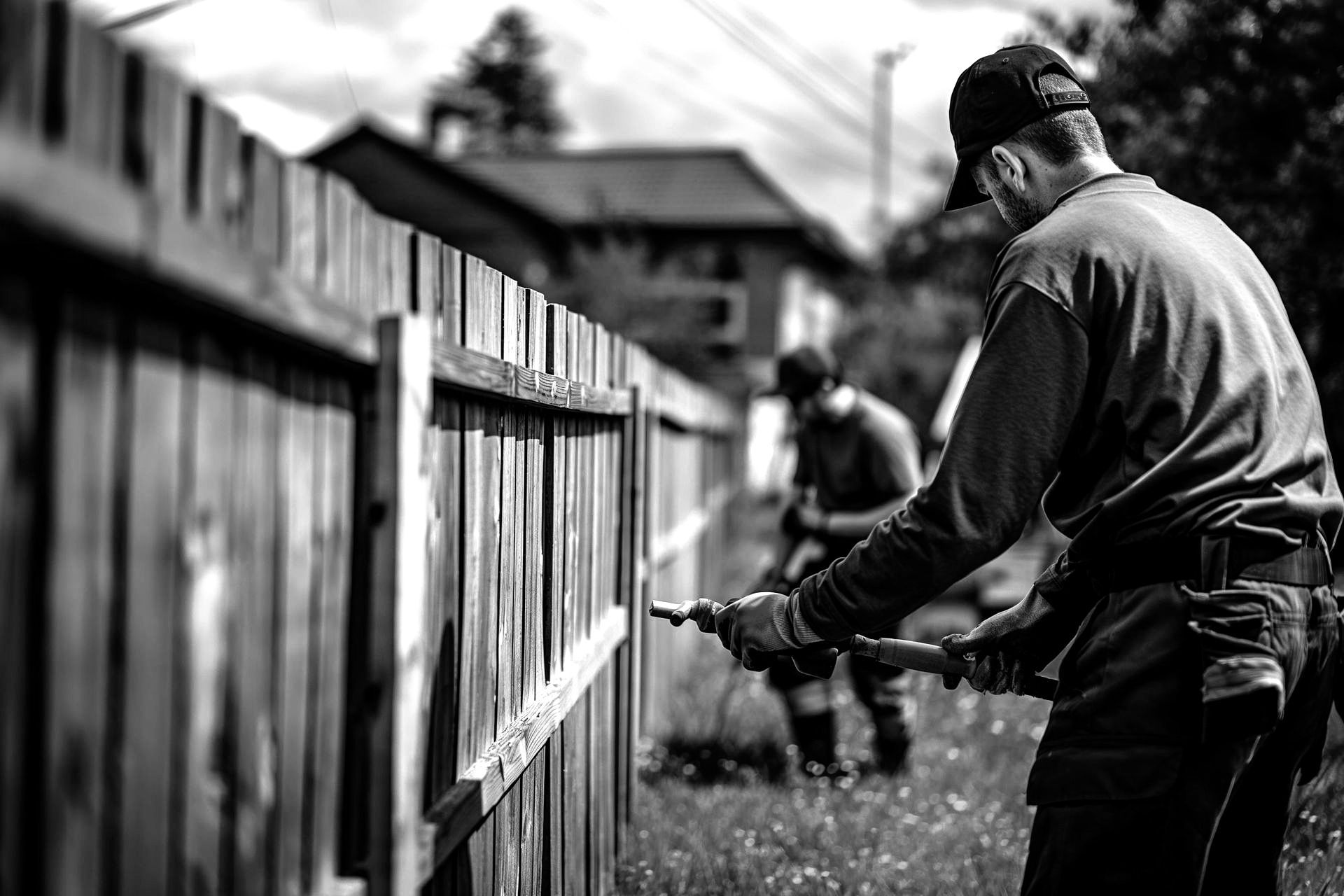Man spraying a wooden fence with another worker in the background. Black and white, outdoors.