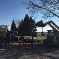 A yellow backhoe loader operating near a chipper machine, trees and bright sky in the background. Construction or cleanup in progress.