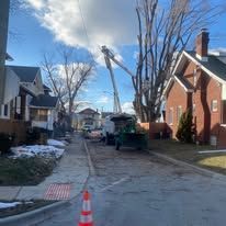 Street scene with a cherry picker, utility truck, and a house with a red brick chimney. A traffic cone sits in the foreground.