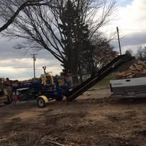 A log splitter with a conveyor belt loading wood into a truck bed in a rural setting.
