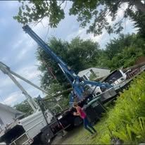 Blue crane truck on a hillside, a person in red shirt next to it. Trees and cloudy sky in the background.