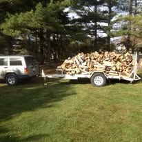 A white SUV pulling a trailer loaded with firewood on a grassy lawn. Sunny day.