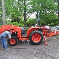 A person working on an orange tractor outdoors. The tractor has a front loader and a rotary cutter.