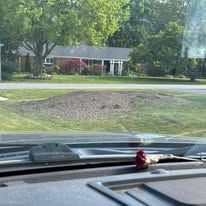View from a car dashboard of a house with a green lawn and a patch of dirt in front of it, with trees in the background.