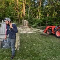 Man in hat holding rake, near tractor and hay, in a grassy outdoor setting with trees.