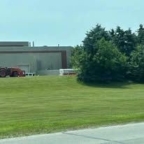 Red tractor and trailer sit on grass in front of a large building and trees on a sunny day.