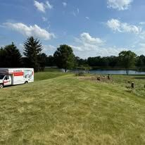 U-Haul truck parked on a grassy hill, with a pond and trees in the background under a blue sky.