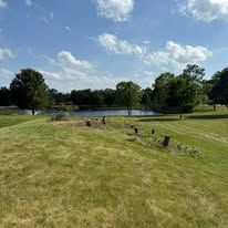 Green field slopes down towards a pond, with trees on the far side. Blue sky with fluffy clouds overhead.
