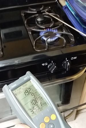 A person is holding a digital thermometer in front of a gas stove.
