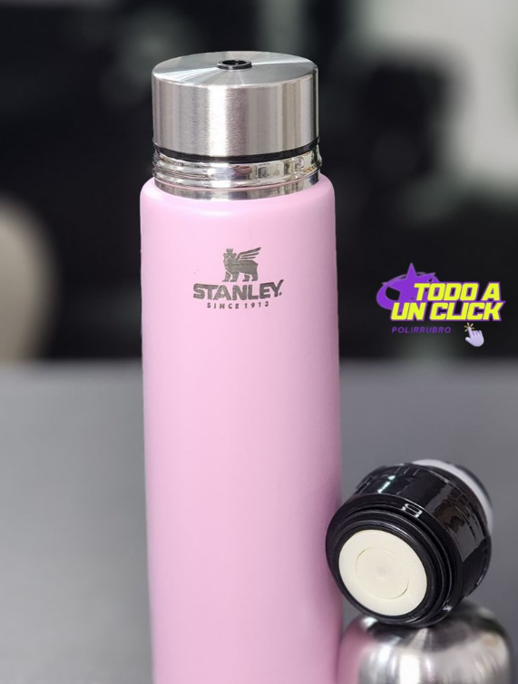 Botella térmica Stanley de color rosa con tapa plateada y tapón negro independiente, sobre superficie reflectante.