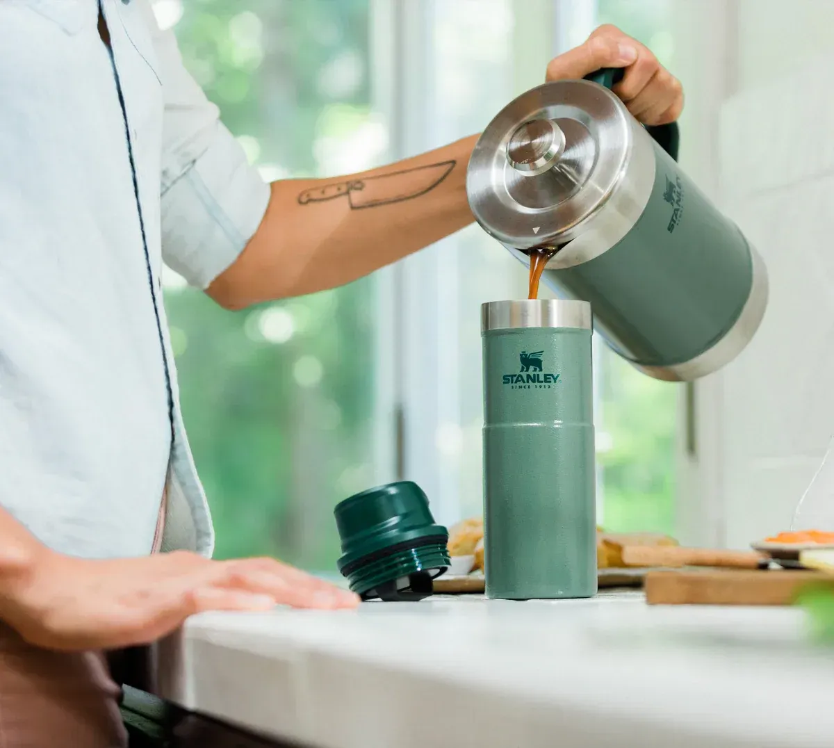 Persona vertiendo café de una prensa francesa Stanley verde en una taza de viaje verde.