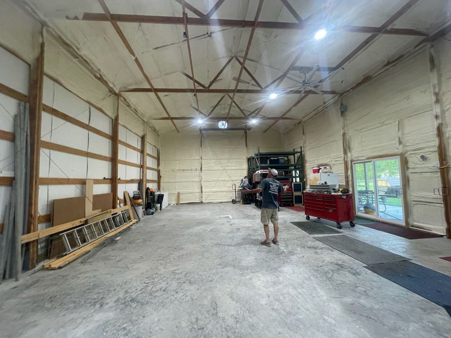 Man in garage with spray foam insulation on walls. Concrete floor, open door, tools, and overhead lights.