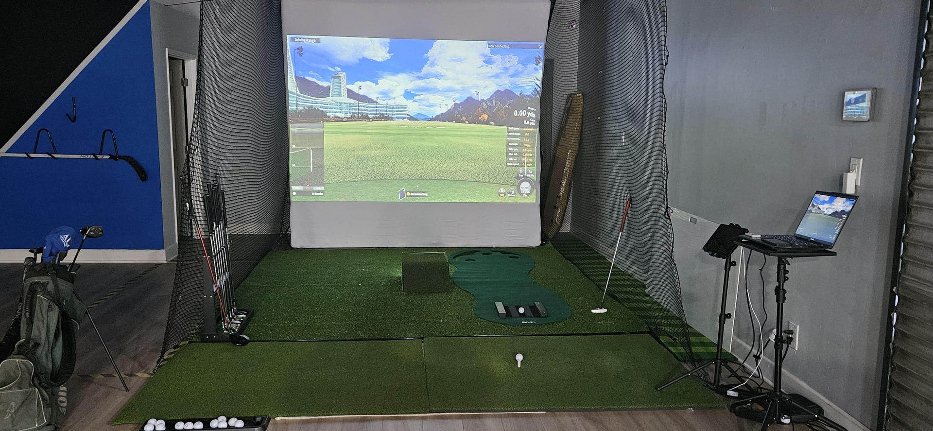 A room with a golf simulator and a laptop on a table.