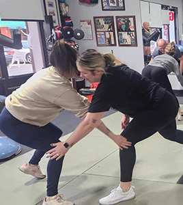 A woman is helping another woman do squats in a gym.