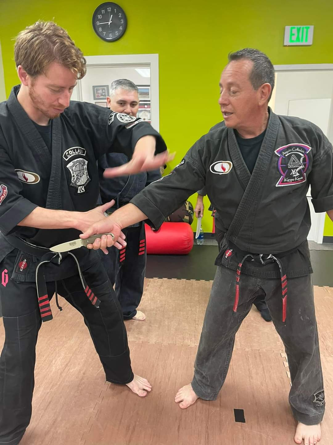 Two men are practicing martial arts in a gym with an exit sign in the background