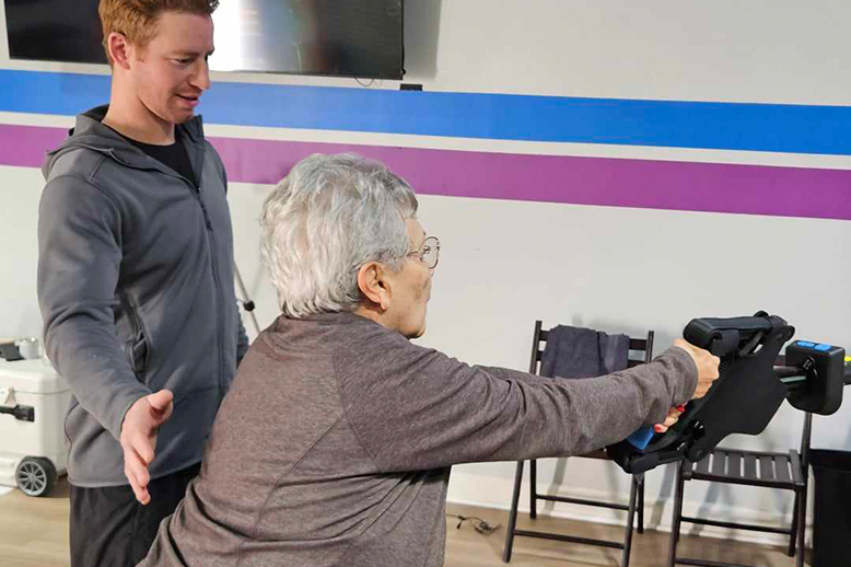 A man is helping an elderly woman with a exercise machine.