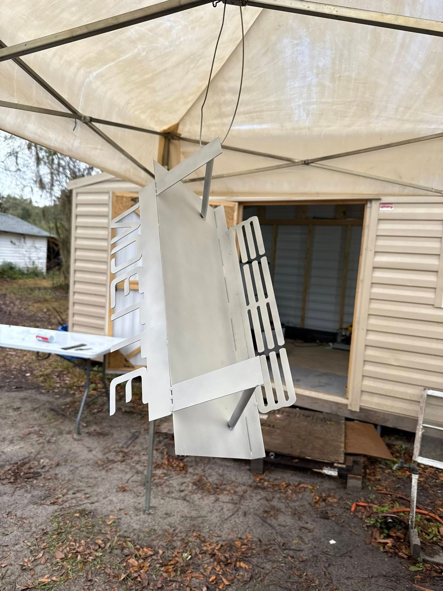 Shelving unit partially assembled outside a tan shed, under a white canopy.