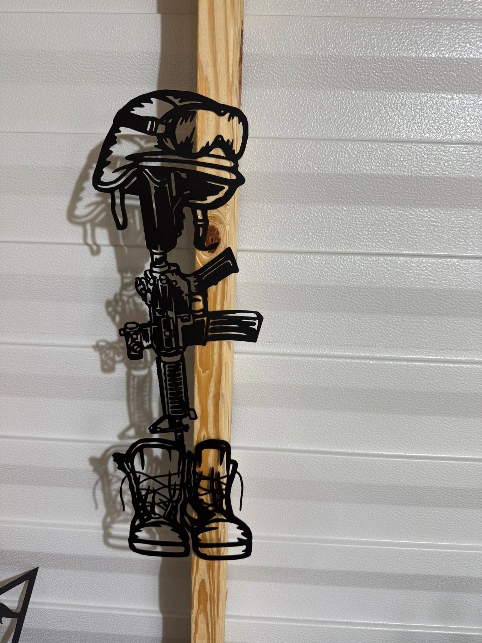 Black metal artwork of a fallen soldier's helmet, rifle, and boots, mounted on wood.