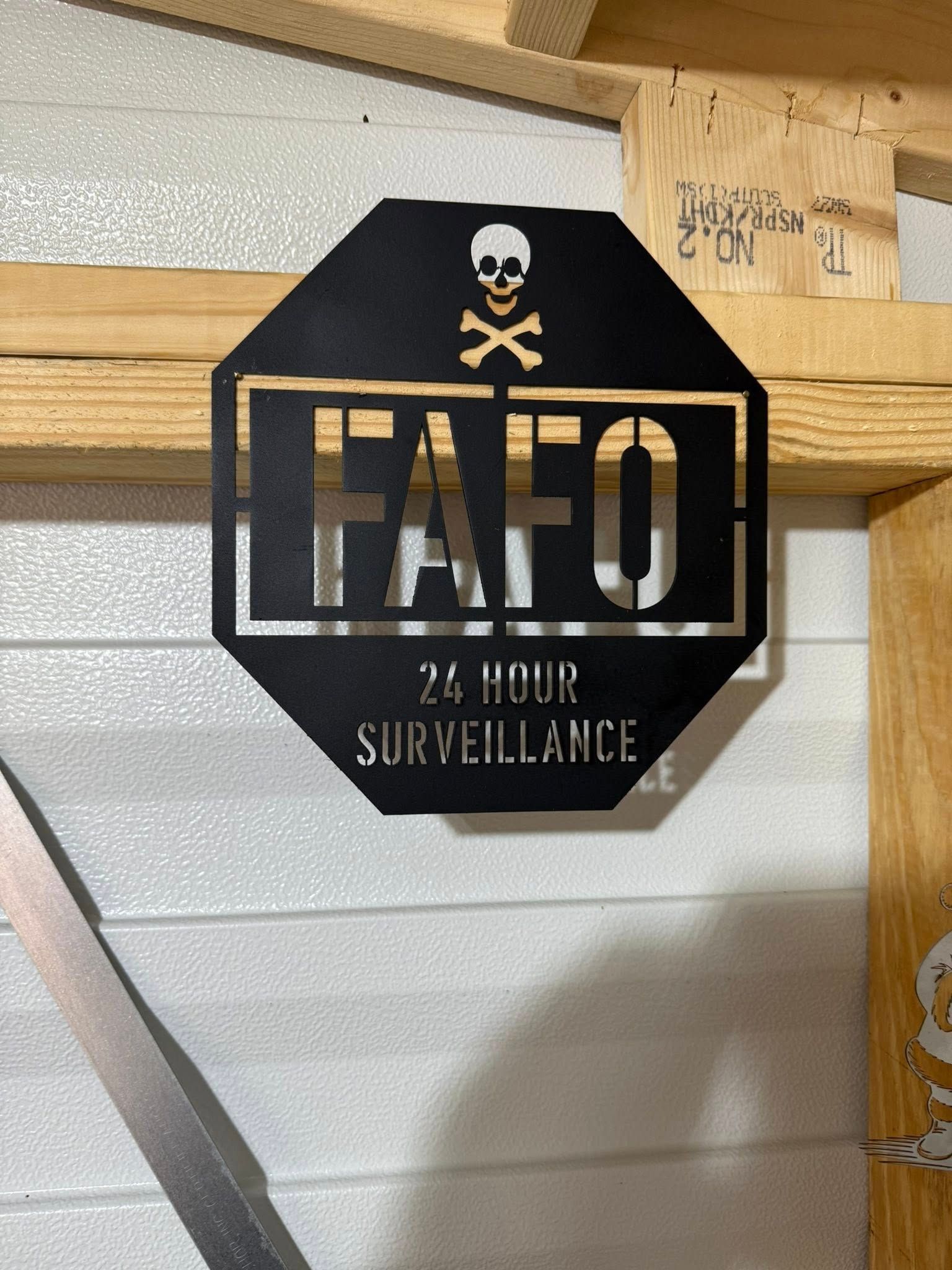 Black metal sign: FAFO, 24-hour surveillance, skull and crossbones. Mounted on wood frame.