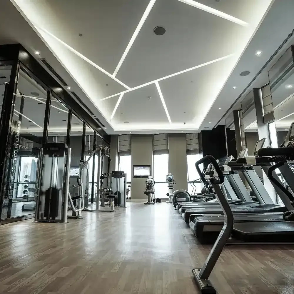 Modern gym with treadmills, weight machines, and large windows. The ceiling has geometric lighting.