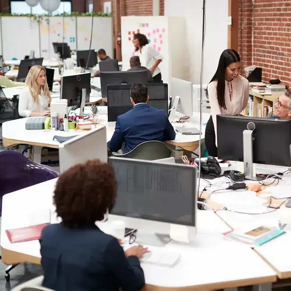 Open-plan office with people at desks, working on computers, and talking to each other.