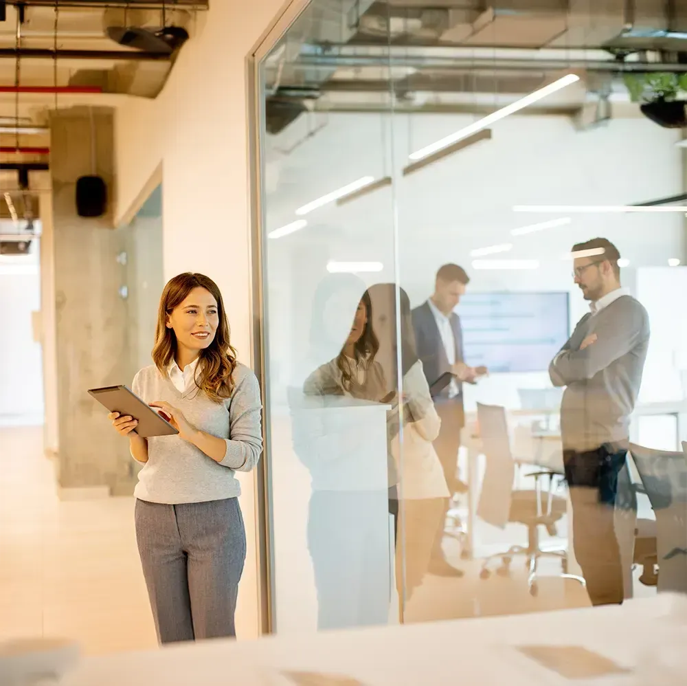 Woman in office corridor holding tablet, looking toward meeting room with people.