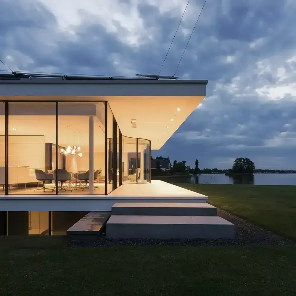 Modern home with glass walls, steps leading to entrance, overlooking a lake under a cloudy sky.