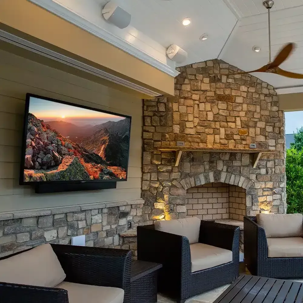 Outdoor entertainment area: TV, fireplace, seating. Beige and stone walls, wooden ceiling, landscape on screen.