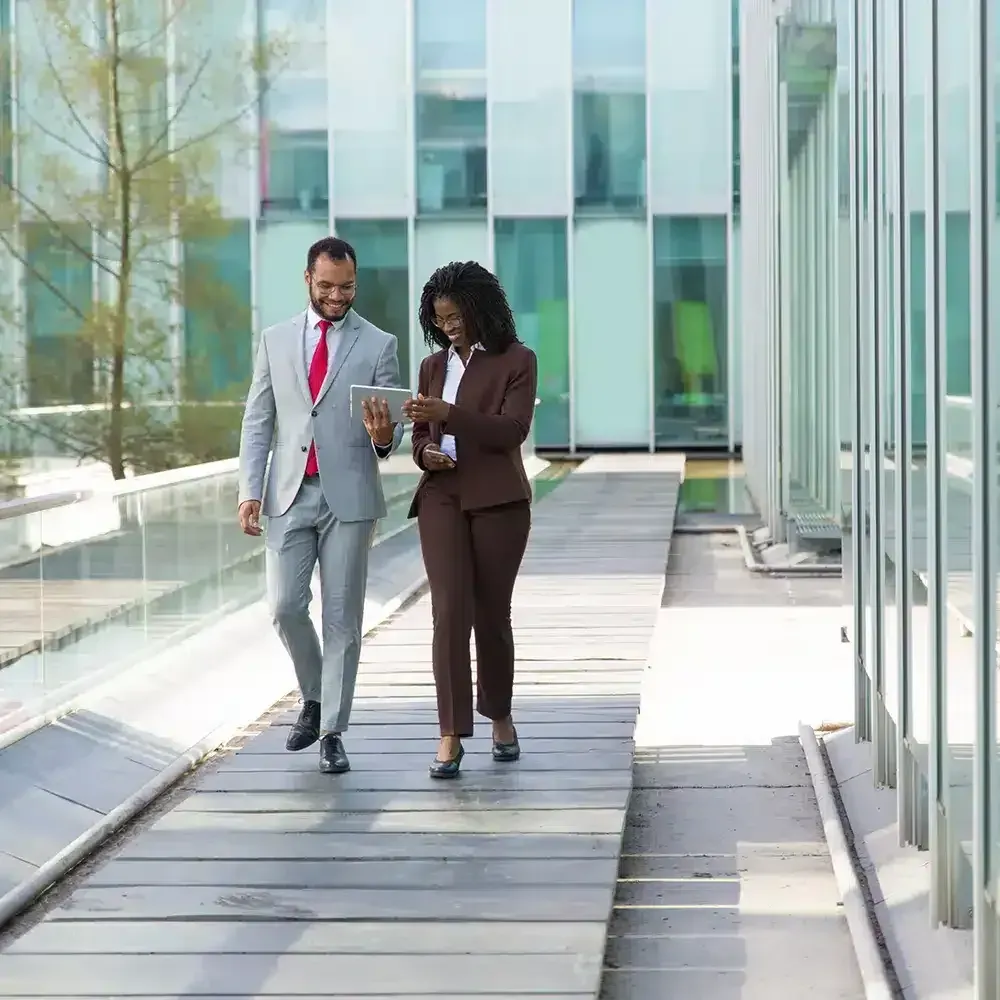 Two professionals in business attire walk and look at a tablet together outdoors. Modern glass building.