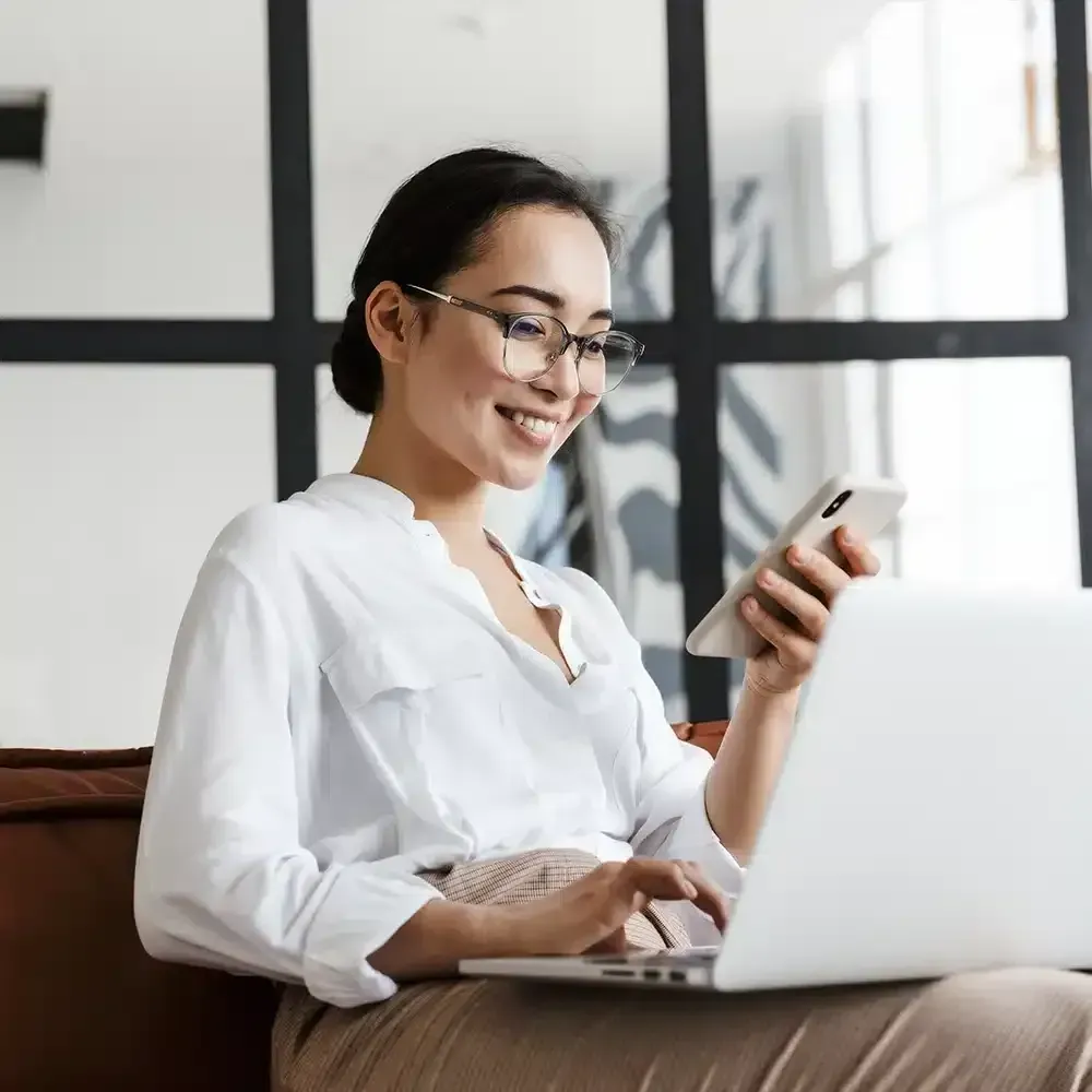 Woman with glasses smiling, using a laptop and phone, in a brightly lit room.