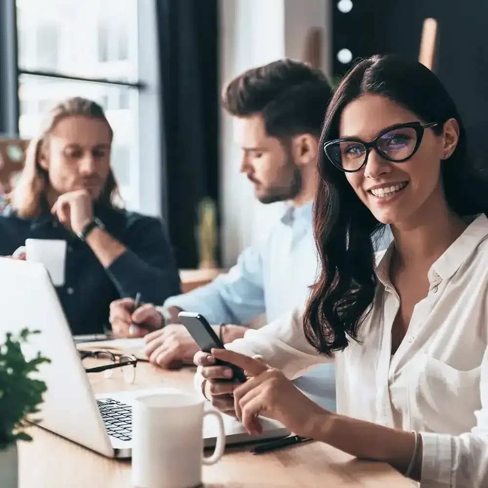 Woman with glasses smiling, holding a phone, sitting at a table with a laptop, two colleagues in the background.