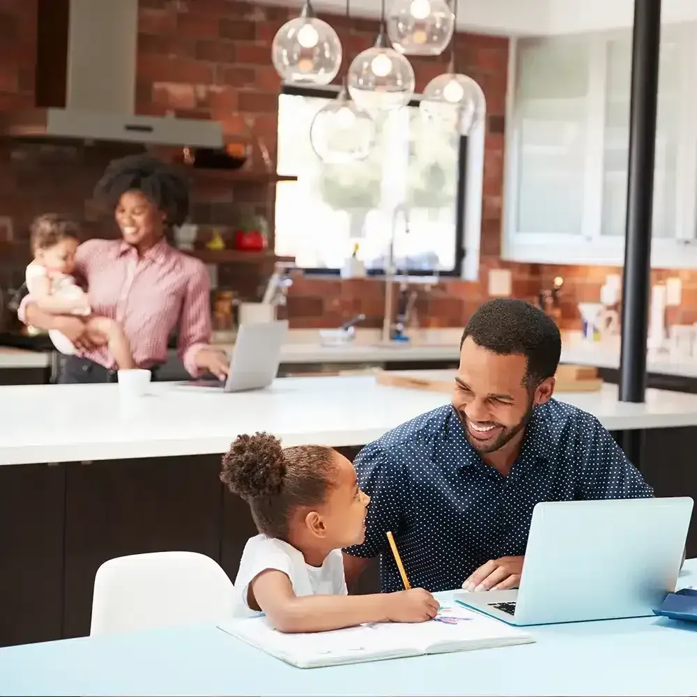 Family in kitchen: Father and daughter at table with laptop and notebook; mother with baby in background.