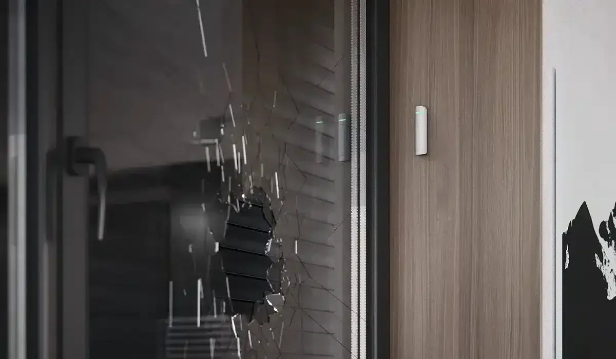 Shattered glass door panel, with a hole and cracks. Adjacent wood-paneled wall and a white sensor are visible.